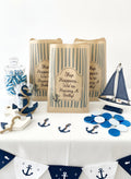 Nautical-themed baby shower party decorations and table setting, featuring brown paper bags with blue stripes, a glass jar of blue and white candy, a wooden anchor, a sailboat, blue and white round confetti, and a banner with anchor prints on a white tablecloth. Ship happens were having a baby 