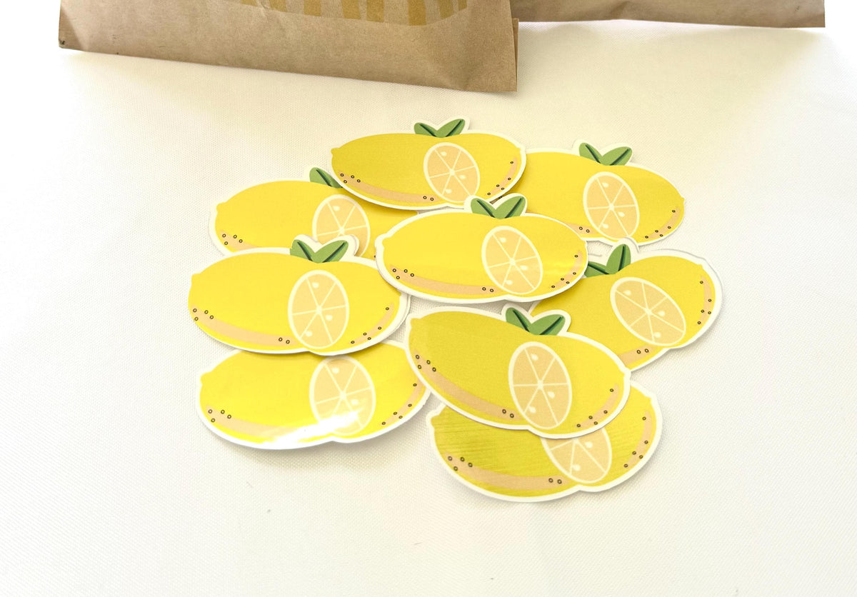A pile of die-cut yellow lemon stickers with green leaves, displayed on a white surface with a brown paper bag partially visible in the background.