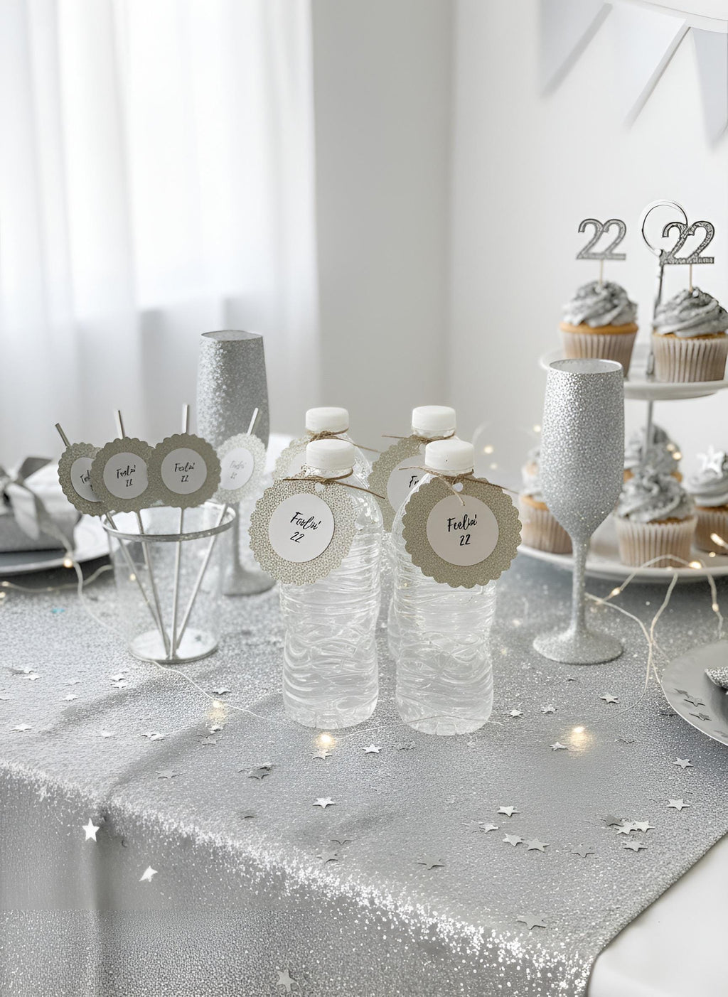 Silver glitter party table decorations including water bottles with place tags, silver cups, a glittery wine glass, and cupcakes on a tiered stand in a bright white room. Gold, silver, black, rose gold glitter favor tags reading feeling 22