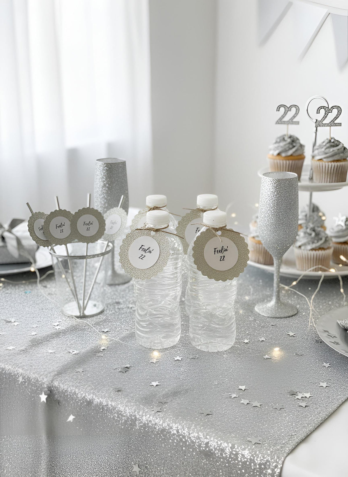Silver glitter party table decorations including water bottles with place tags, silver cups, a glittery wine glass, and cupcakes on a tiered stand in a bright white room. Gold, silver, black, rose gold glitter favor tags reading feeling 22