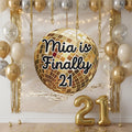 A gold disco ball balloon with the text 'Finally 21' in black script, displayed against a decorated party backdrop with gold and silver balloons, gold tinsel, string lights, and gold number 21 foil balloons on the floor.