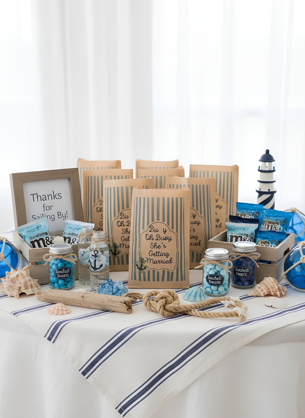 A nautical-themed bridal shower gift table display featuring striped paper bags, candy jars a lighthouse decoration, and seashells on a white tablecloth with blue stripes. Buoy oh buoy she’s getting married