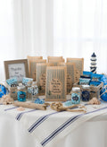 A nautical-themed bridal shower gift table display featuring striped paper bags, candy jars a lighthouse decoration, and seashells on a white tablecloth with blue stripes. Buoy oh buoy she’s getting married