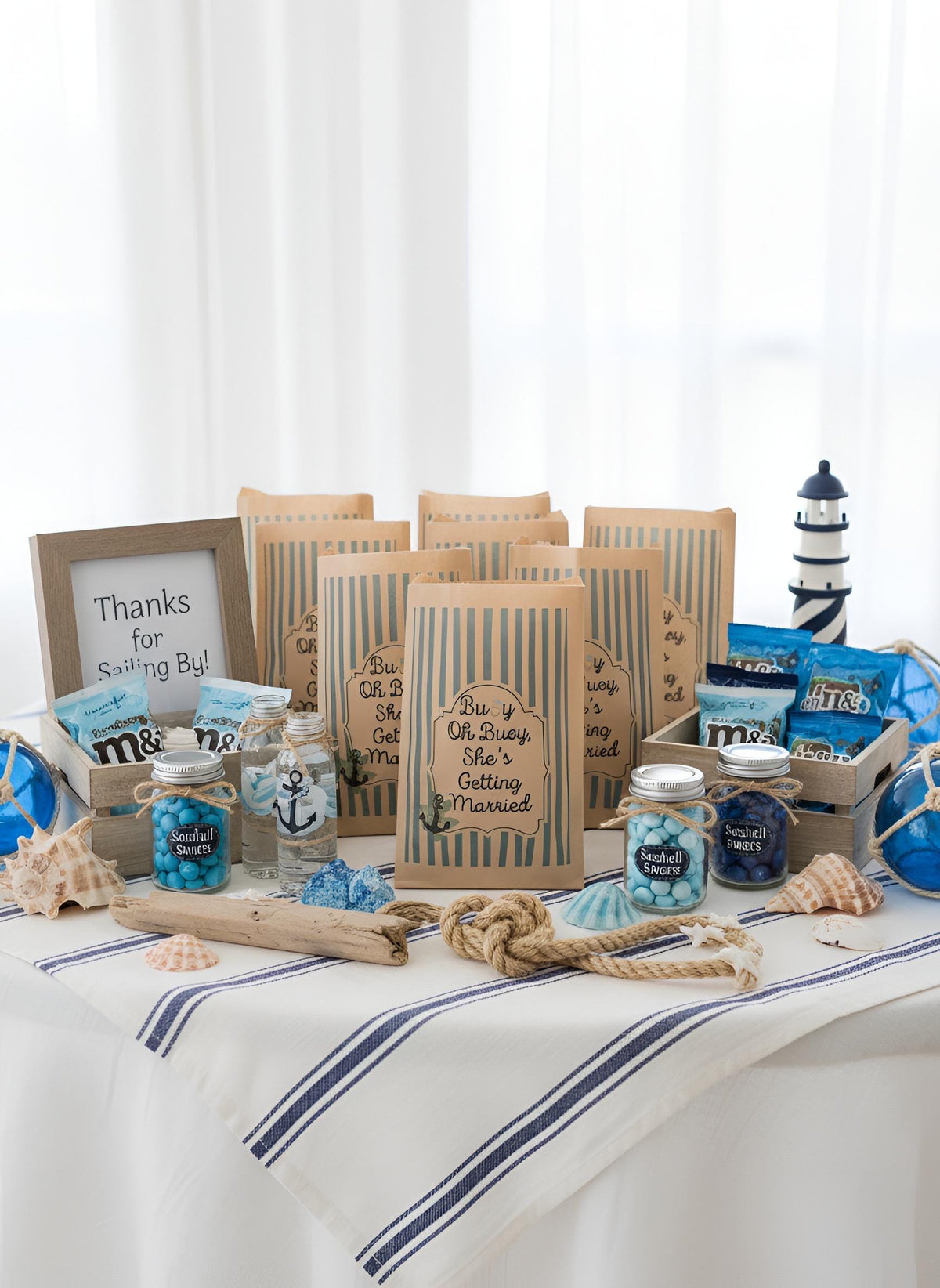 A nautical-themed bridal shower gift table display featuring striped paper bags, candy jars a lighthouse decoration, and seashells on a white tablecloth with blue stripes. Buoy oh buoy she’s getting married