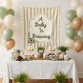 A styled baby shower party setup with a white tablecloth-covered table, pastries, plants, and a striped backdrop featuring the phrase 'A Baby Is Brewing' in a decorative sign. Balloons in white, sage green, and blush pink frame the scene, with warm string lights adding a cozy glow.