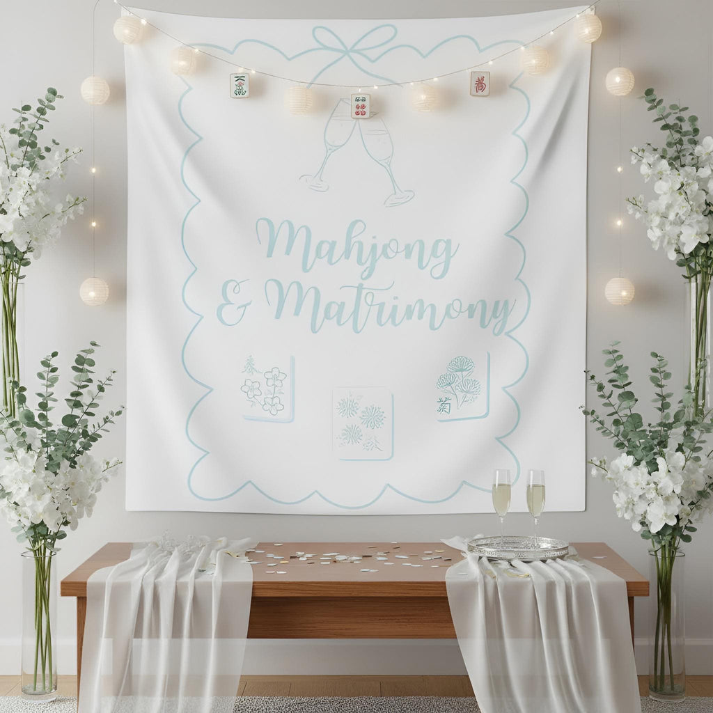 A large white fabric tapestry with light blue decorative border and the words 'Mahjong & Matrimony' hangs on a wall above a wooden table with draped white fabric and two champagne glasses. The scene is styled with string lights, tall floral arrangements, and a clean indoor setting.