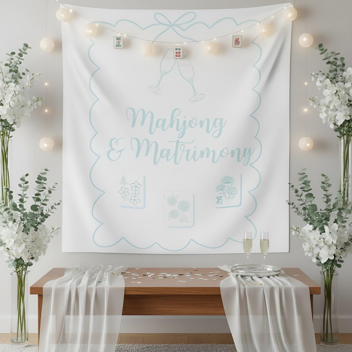 A large white fabric tapestry with light blue decorative border and the words 'Mahjong & Matrimony' hangs on a wall above a wooden table with draped white fabric and two champagne glasses. The scene is styled with string lights, tall floral arrangements, and a clean indoor setting.