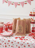 Strawberry-themed baby shower party supplies arranged on a table, including paper bags with printed labels, red-and-white polka dot plates, cups, jars of candy, and a matching tablecloth. A cupcake stand with frosted cupcakes is visible in the background, along with a red-and-white gingham banner and cherry bunting on the wall. A berry cute baby is on the way