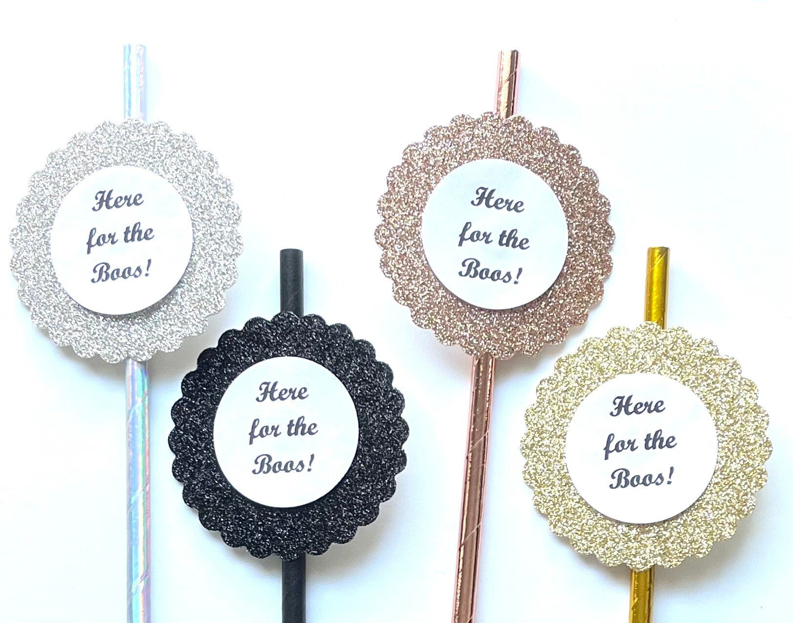 Four glitter paper straws, each with a scalloped circular glitter frame and a white center label reading 'Here for the Boos!'. The toppers come in silver, black, rose gold, and gold colors, arranged on a plain white background.