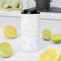 A white insulated can cooler with a black lid, featuring light blue printed champagne glasses and the text 'Mahjong & Mimosas', set on a white marble countertop surrounded by fresh lemons and limes with a blurred kitchen background.