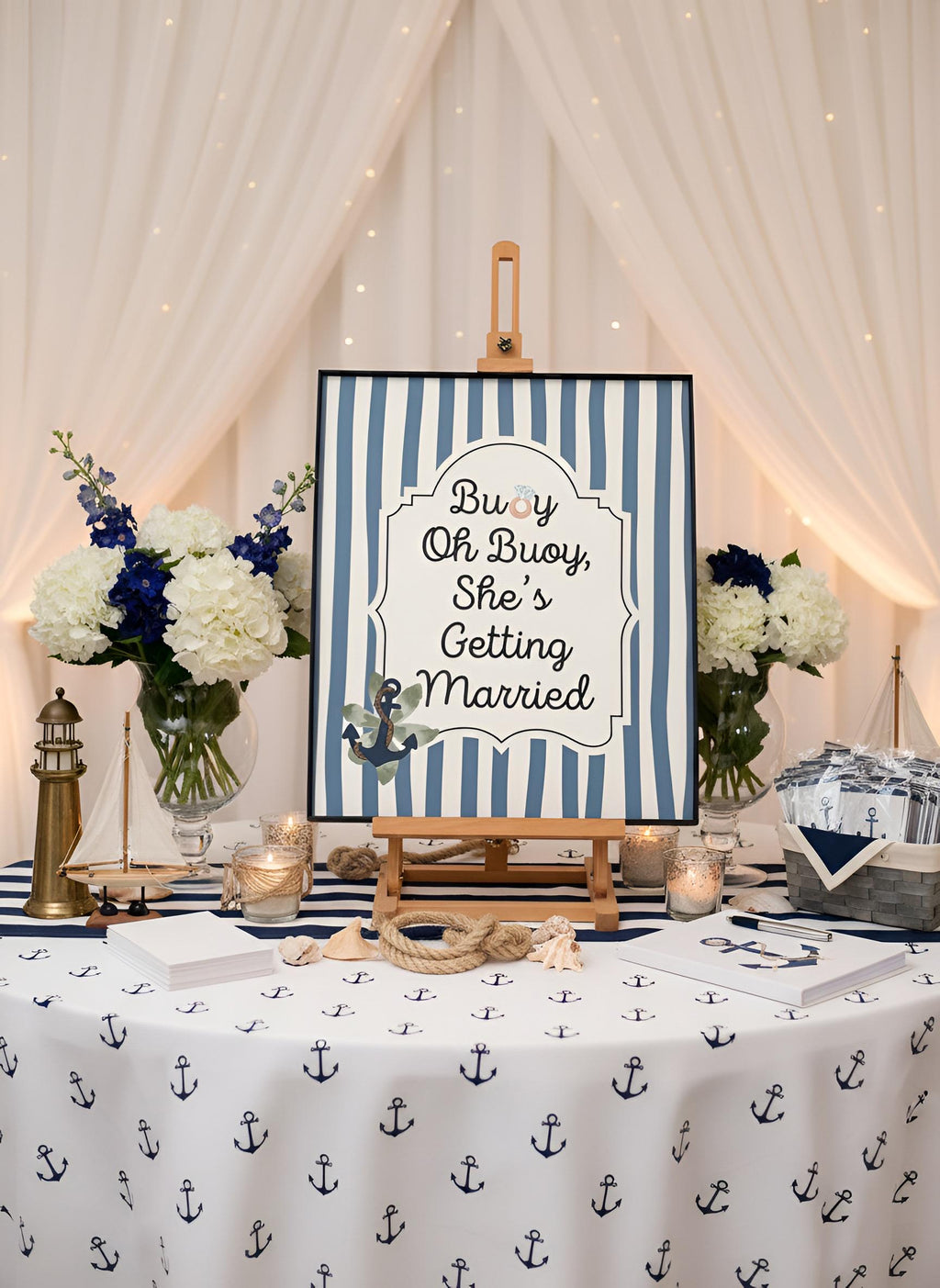 A nautical-themed wedding sign on a wooden easel that reads 'Buoy Oh Buoy She's Getting Married' with blue and white stripes and an anchor illustration. The sign is displayed on a decorated table with an anchor-patterned tablecloth, floral arrangements, candles, and nautical decor against a backdrop of draped white fabric and string lights.