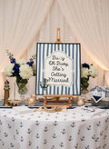 A nautical-themed wedding sign on a wooden easel that reads 'Buoy Oh Buoy She's Getting Married' with blue and white stripes and an anchor illustration. The sign is displayed on a decorated table with an anchor-patterned tablecloth, floral arrangements, candles, and nautical decor against a backdrop of draped white fabric and string lights.