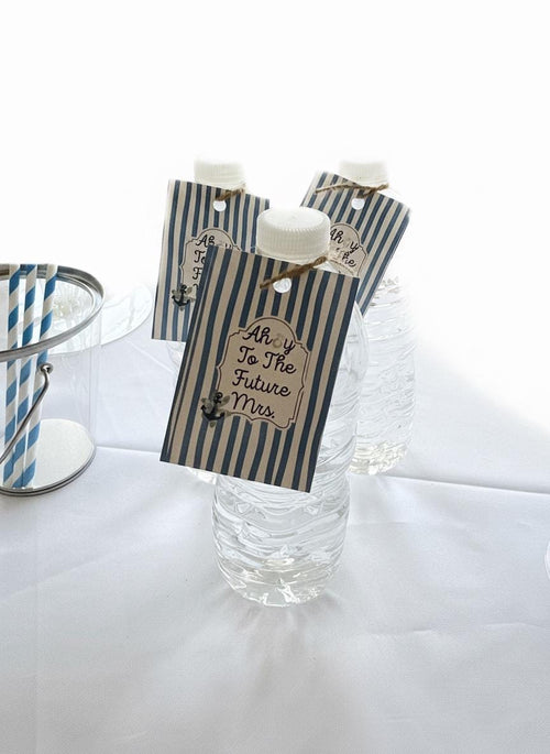 Three clear plastic water bottles with white caps, each decorated with a navy and beige striped paper tag that reads 'AhoY To The Future Mrs.' and tied with twine, set on a white draped surface.