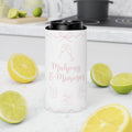 A white insulated can cooler sleeve with pink champagne-themed graphics and the text 'Mahjong & Mimosas' stands on a white kitchen counter surrounded by sliced lemons and limes.