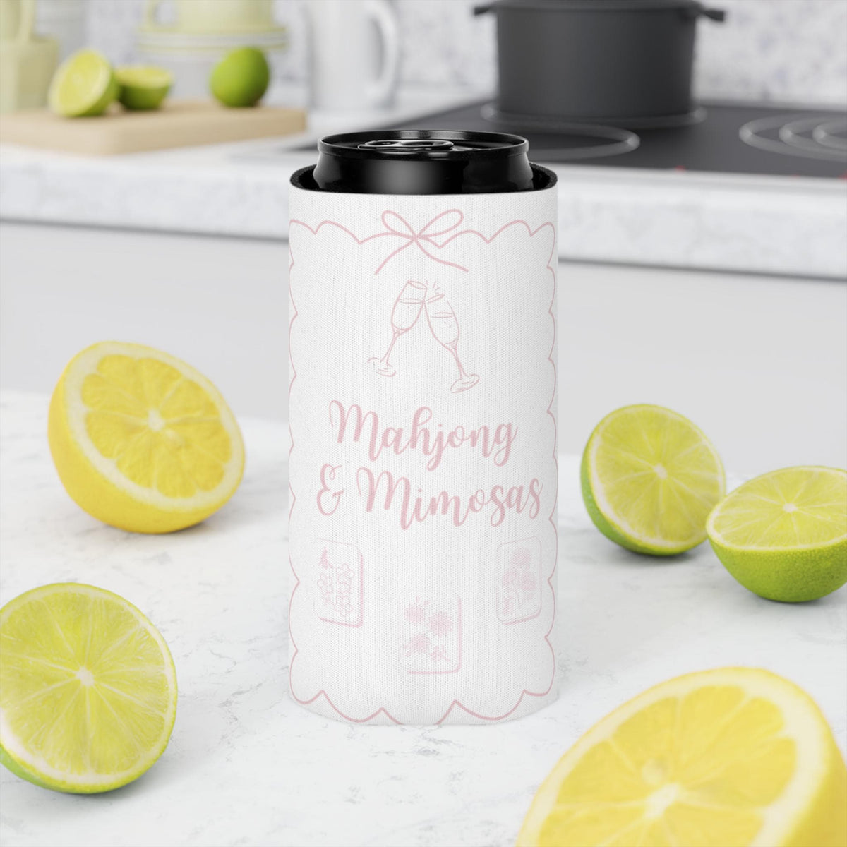A white insulated can cooler sleeve with pink champagne-themed graphics and the text 'Mahjong & Mimosas' stands on a white kitchen counter surrounded by sliced lemons and limes.