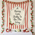 A strawberry-themed baby shower backdrop tapestry with red and cream vertical stripes, a central cream label reading 'A Berry Cute Baby Is On The Way,' and decorative burlap 'BABY SHOWER' banners above. Strawberry garlands and a styled table with strawberries, candles, and flowers are arranged in front.