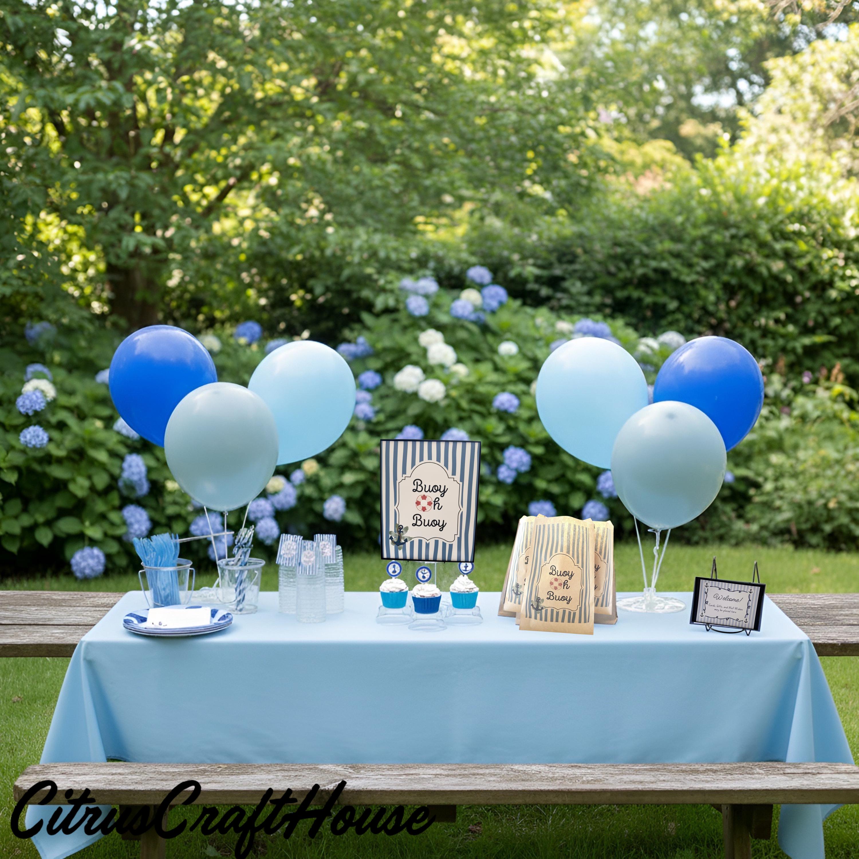 A baby shower party setup on an outdoor wooden bench with a light blue tablecloth, blue and pale blue balloons, baby books, and small blue cupcakes, set against a green garden background with hydrangeas. Buoy oh Buoy