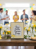 A square sign reading 'Our Little Lemon Drop' with yellow and white stripes and lemon illustrations, displayed on a wooden table in a party setting with blurred people in the background.