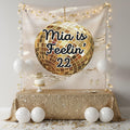 A personalized party backdrop setup featuring a large gold disco ball graphic with the text 'Feelin' 22' on a champagne-colored tapestry. The scene is decorated with a gold sequin tablecloth, white balloons, string lights, and a small cake on a table.