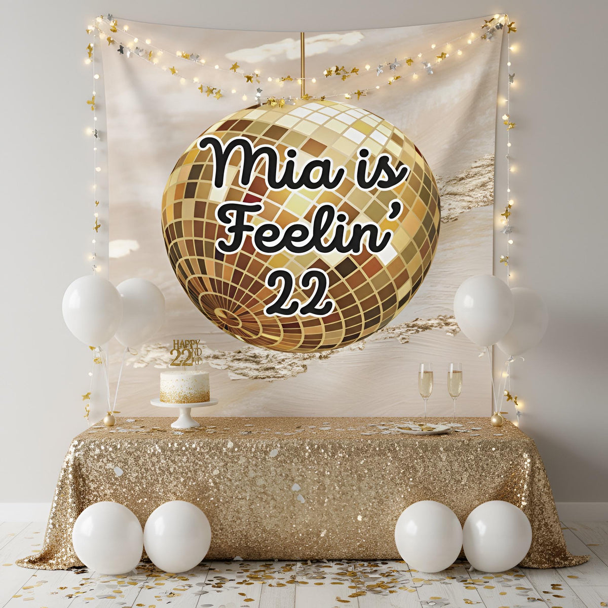 A personalized party backdrop setup featuring a large gold disco ball graphic with the text 'Feelin' 22' on a champagne-colored tapestry. The scene is decorated with a gold sequin tablecloth, white balloons, string lights, and a small cake on a table.