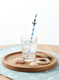 A clear glass tumbler filled with a clear bubbly drink sits on a round wooden tray, with a blue-and-white striped paper straw featuring a small black anchor decoration. The tray is styled with seashells, a rope coil, and driftwood on a light blue patterned cloth against a white background.