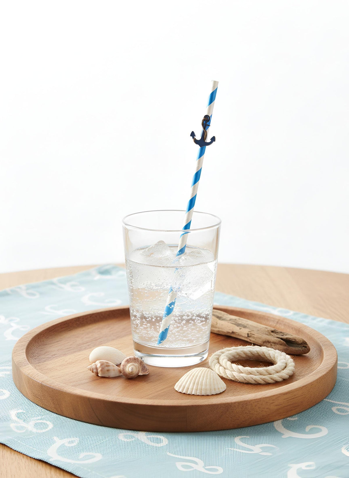 A clear glass tumbler filled with a clear bubbly drink sits on a round wooden tray, with a blue-and-white striped paper straw featuring a small black anchor decoration. The tray is styled with seashells, a rope coil, and driftwood on a light blue patterned cloth against a white background.