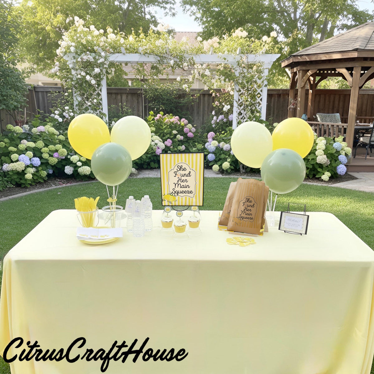 A styled outdoor party table setup in a garden with a pale yellow lemon tablecloth, yellow and mint balloons, paper cups, napkins, and a sign reading 'She found her main squeeze.' A wooden gazebo and flowering plants are visible in the background.