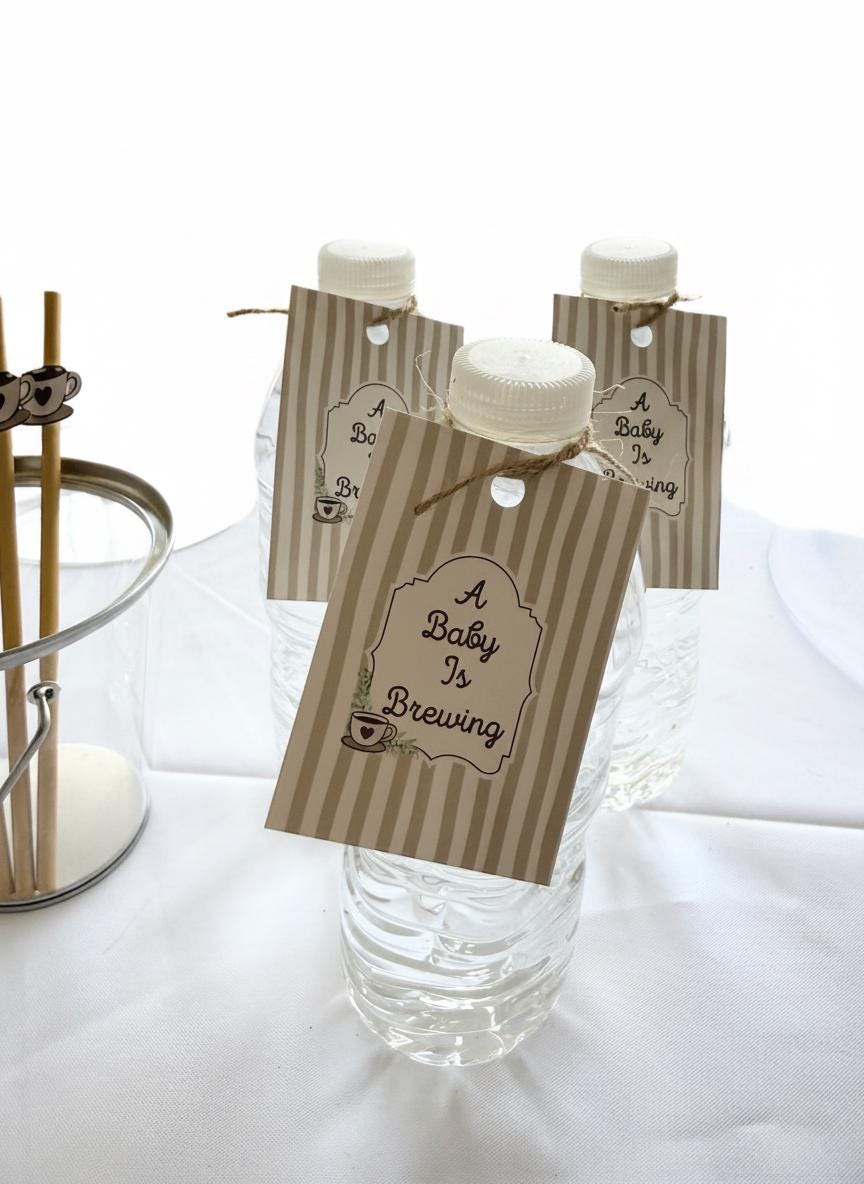 Three clear plastic water bottles with white caps, each decorated with a beige and brown striped paper tag that reads 'A Baby Is Brewing,' arranged on a white tablecloth.