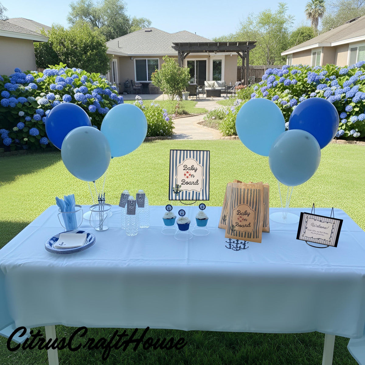 A baby shower party setup in a backyard lawn, featuring a table covered with a light blue tablecloth, blue and white plates, napkins, cupcakes, a brown paper bag, and matching blue and white balloons. The background shows a suburban house, greenery, and blue hydrangeas.  Baby on board
