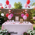 A styled outdoor birthday party setup with a table covered in a pink tablecloth, decorated with red, pink, and white balloons, a pink-and-white striped sign, balloons, banners, cups, and flowers in a backyard setting. the cherry on top for the bride