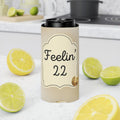 A beige insulated can cooler sleeve with a black lid, printed with the text 'Feelin' 22' and a small gold-toned graphic, standing on a white kitchen counter surrounded by sliced lemons and limes.