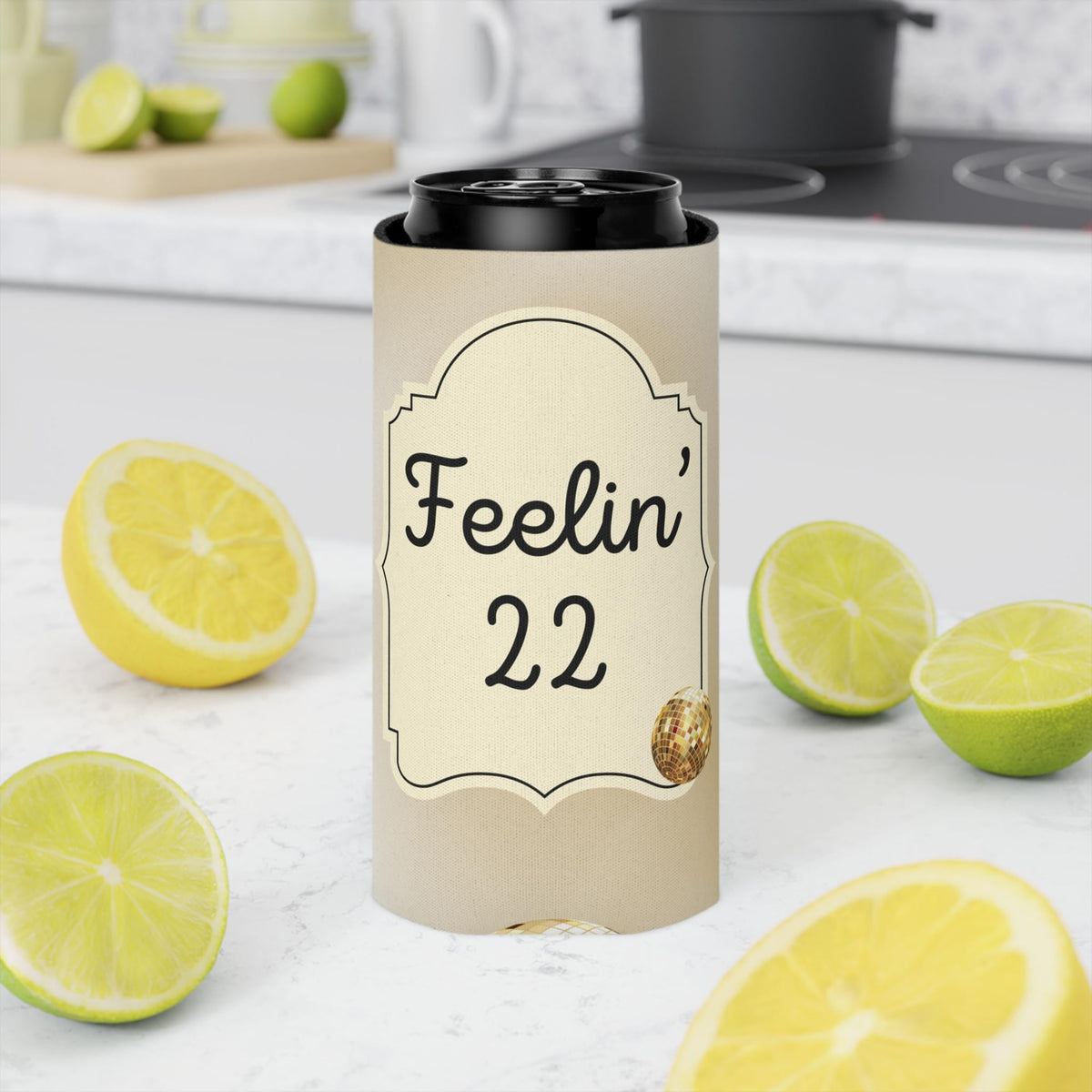 A beige insulated can cooler sleeve with a black lid, printed with the text 'Feelin' 22' and a small gold-toned graphic, standing on a white kitchen counter surrounded by sliced lemons and limes.