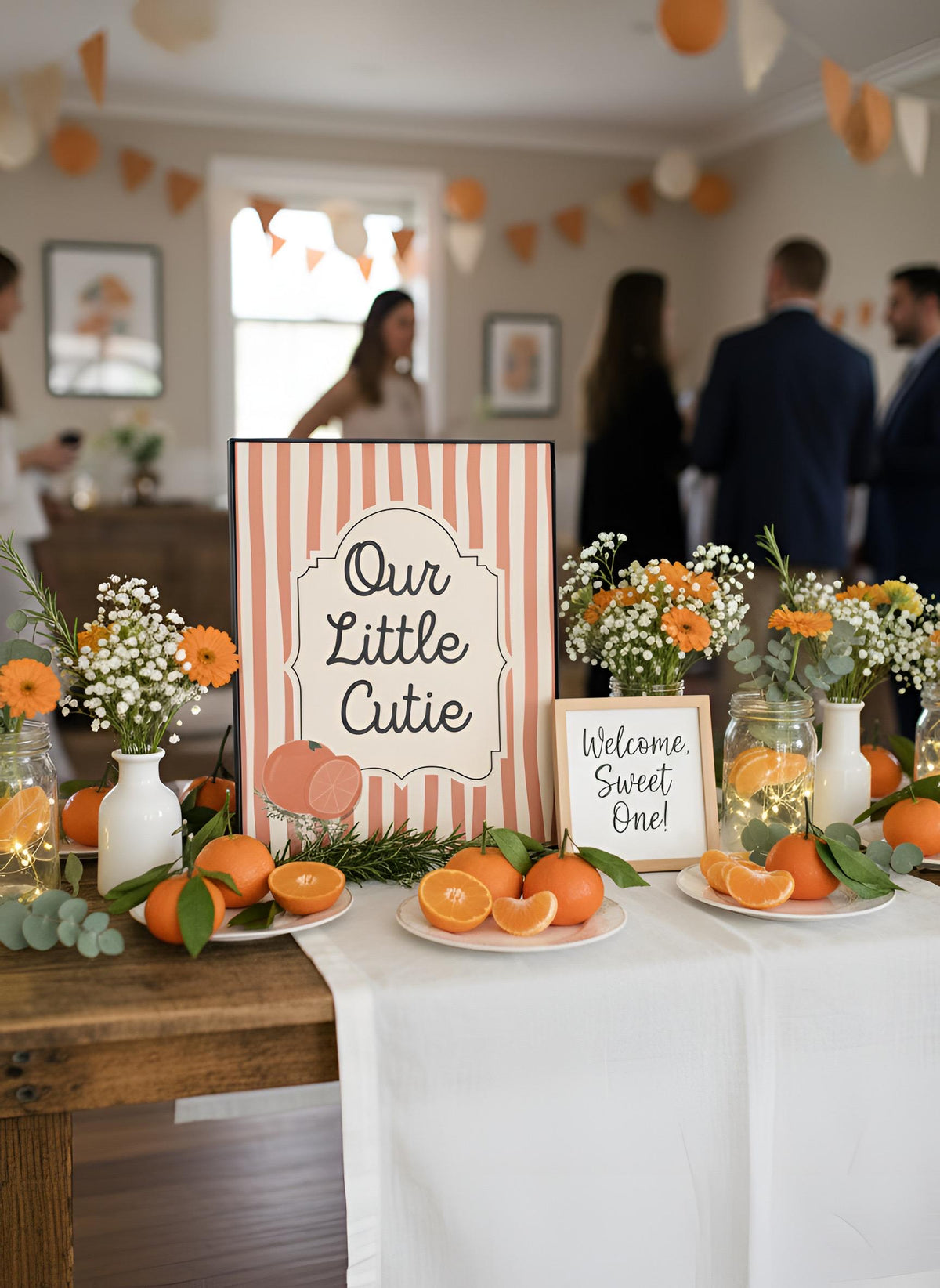 A styled reception table with orange and white theme decorations, including a sign reading 'Our Little Cutie' and another reading 'Welcome Sweet One!', surrounded by oranges, flowers, and greenery, with blurred guests in the background.