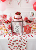 A styled baby shower or bachelorette party dessert table decorated with strawberries, pink and red balloons, cherry-patterned table runners, cupcakes, jars of candy, and a central sign that reads, 'A Berry Cute Baby Is On The Way.'.