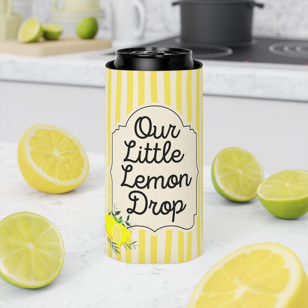 A yellow and white striped can cooler sleeve with the text 'Our Little Lemon Drop' stands on a white countertop, surrounded by fresh lemon slices and limes in a kitchen setting.