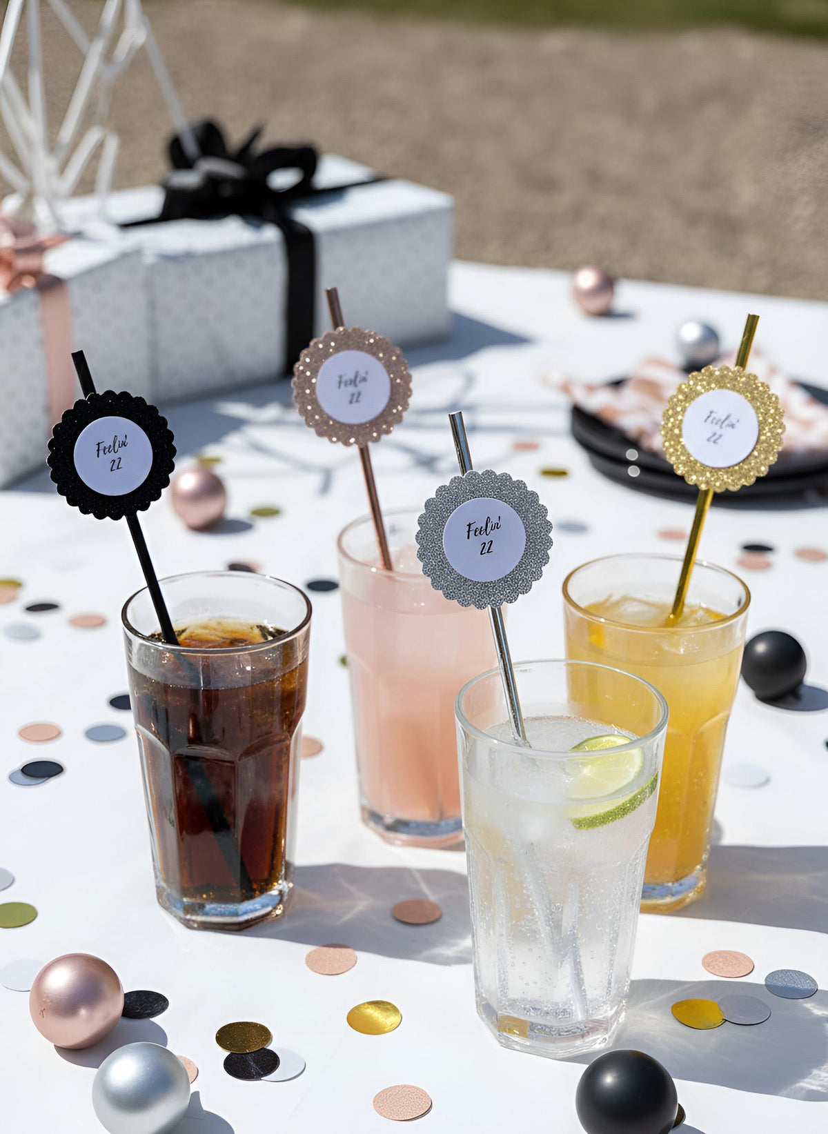 Four decorated cocktail glasses with straws on a white tablecloth outdoors. The glasses contain Rose gold, gold, silver, black glitter, decorations, saying feeling 22