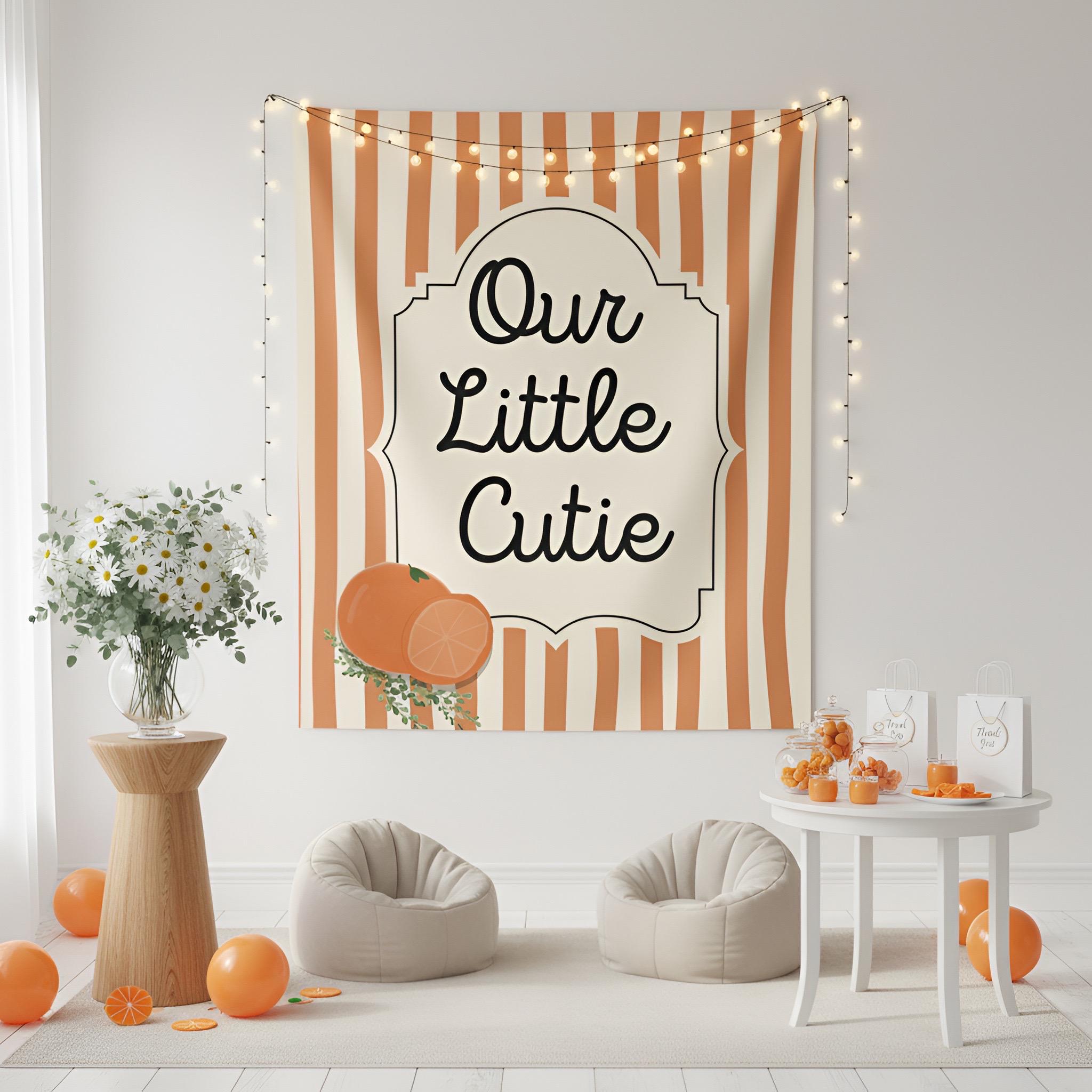 A decorative wall tapestry with vertical orange and cream stripes, a cream central label reading “Our Little Cutie” in black script, and orange fruit illustrations, hanging in a styled room with string lights, bean bags, and small orange decor items.