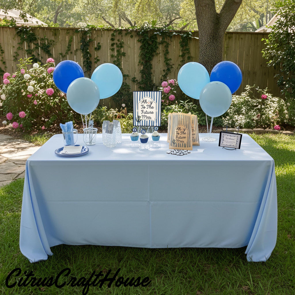 A decorated outdoor party table setup in a backyard garden, featuring a light blue tablecloth, blue and light blue balloons, paper bags, and party supplies. The scene is set on green grass with a wooden fence and flowering bushes in the background. Ahoy to the future Mrs.
