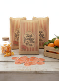 Three kraft paper gift bags with orange vertical stripes and 'Zest Wishes For The Mrs' printed labels, arranged on a white wooden table with scattered orange circular stickers, a glass jar of orange candies, and oranges in a wooden crate.