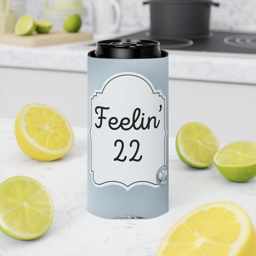 A light blue can cooler sleeve with a white decorative label reading 'Feelin' 22' in black script, shown around a black can on a kitchen counter with lemons and limes around it.