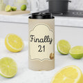 A tall insulated tumbler with a black lid stands on a white countertop in a kitchen setting. The tumbler sleeve is beige with a decorative label that reads 'Finally 21' in black script and numerals, with a small gold glitter accent.