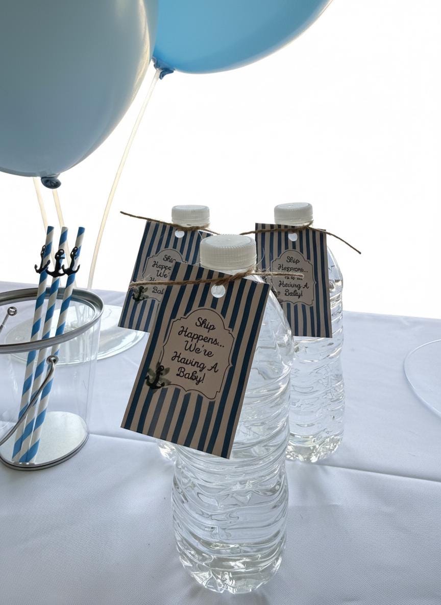 Three clear plastic water bottles with white caps, decorated with navy and white striped paper tags that read 'Ship Happens We're Having A Baby!', set on a white tablecloth with blue balloons and blue-and-white straws in the background.