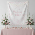 A white fabric backdrop banner with pink decorative borders, champagne glasses, and the words 'Mahjong & Matrimony' in pink script, displayed in a styled event setup with a pink tablecloth, floral arrangements, and small mahjong tiles on a table.