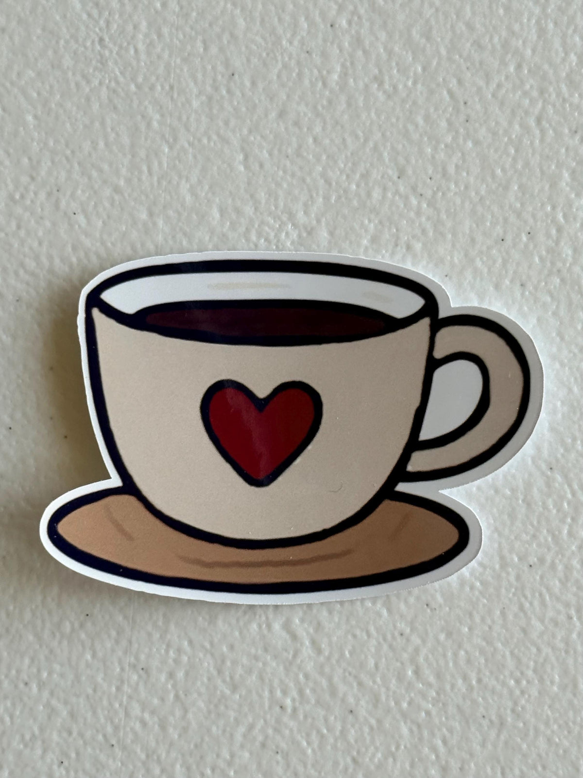 A die-cut sticker of a coffee cup on a saucer with a red heart, shown on a textured off-white background.