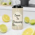 A stainless steel travel tumbler with a black lid sits on a white marble countertop, surrounded by fresh lemon slices and limes. The tumbler has a beige and cream vertical striped sleeve with a central label that reads 'Love Is Brewing' in black script, with a small coffee cup illustration at the bottom.