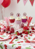 A strawberry-themed party table setting featuring three decorated cupcakes on a glass pedestal stand, red and pink striped candy sticks in jars, cherry-shaped plates, napkins, and utensils, and red cherries scattered on a white tablecloth printed with cherries and strawberries.