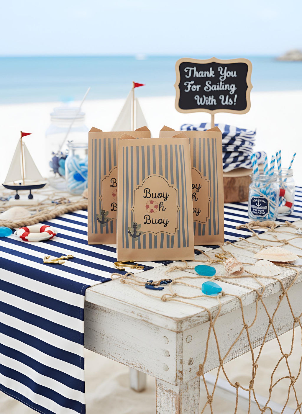 Nautical-themed party favor bags with blue and white stripes and a central sign reading 'Thank You For Sailing With Us!' displayed on a beach-themed table with a striped tablecloth, seashells, rope, and small toy boats. Buoy Oh Buoy