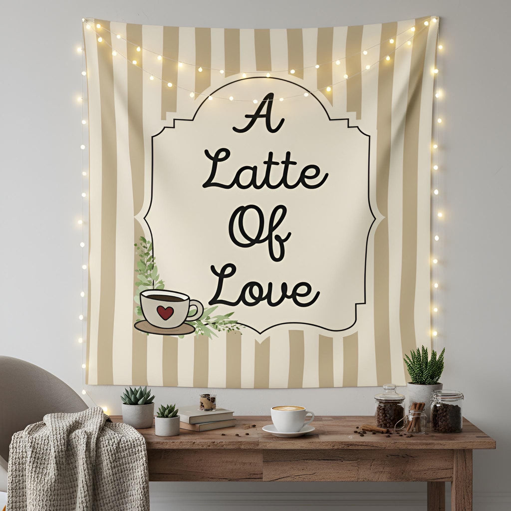 A fabric wall tapestry hanging on a wall above a wooden table, featuring the text 'A Latte Of Love' in black script inside an ornate frame. The tapestry has vertical beige and cream stripes, a small coffee cup illustration with a red heart, and is decorated with warm string lights.