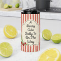 A tall insulated tumbler with a black lid sits on a white countertop in a kitchen setting. The tumbler is wrapped in a red-and-cream striped sleeve with a cream label that reads, "A Berry Cute Baby Is On The Way" and includes small berry illustrations.