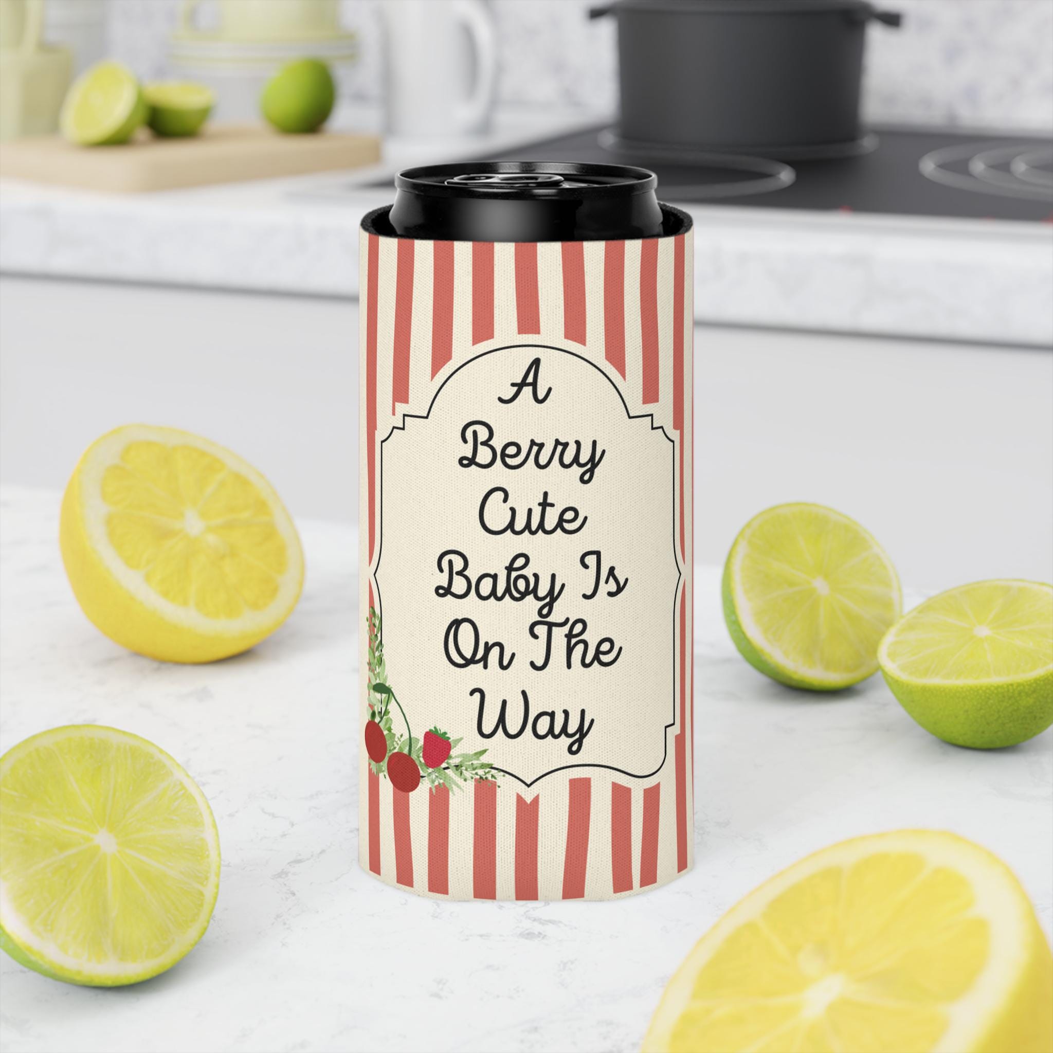 A tall insulated tumbler with a black lid sits on a white countertop in a kitchen setting. The tumbler is wrapped in a red-and-cream striped sleeve with a cream label that reads, "A Berry Cute Baby Is On The Way" and includes small berry illustrations.